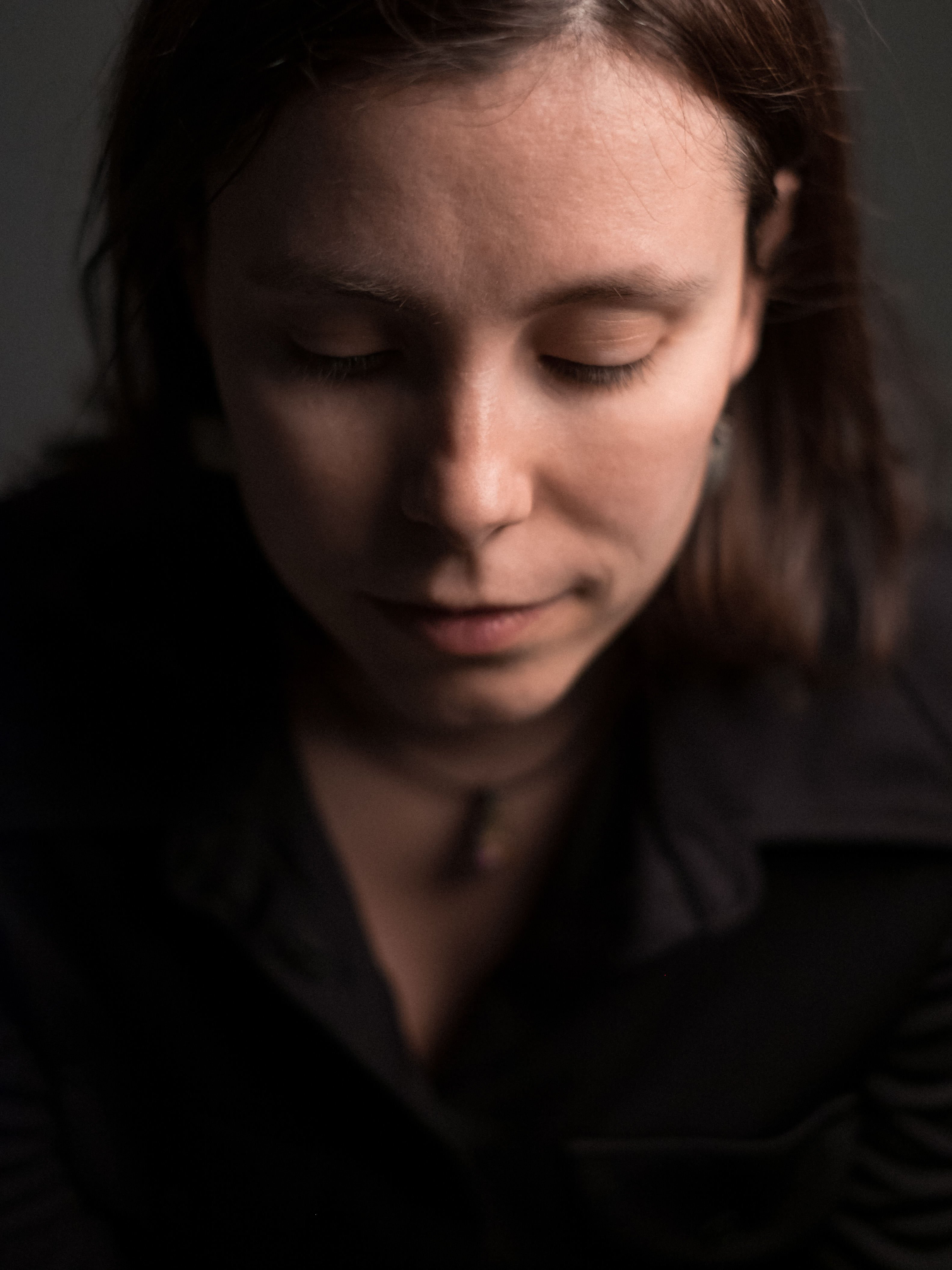 Primo piano verticale del volto con occhi chiusi; capelli corti, collana sottile al collo, luce morbida frontale, sfondo scuro.