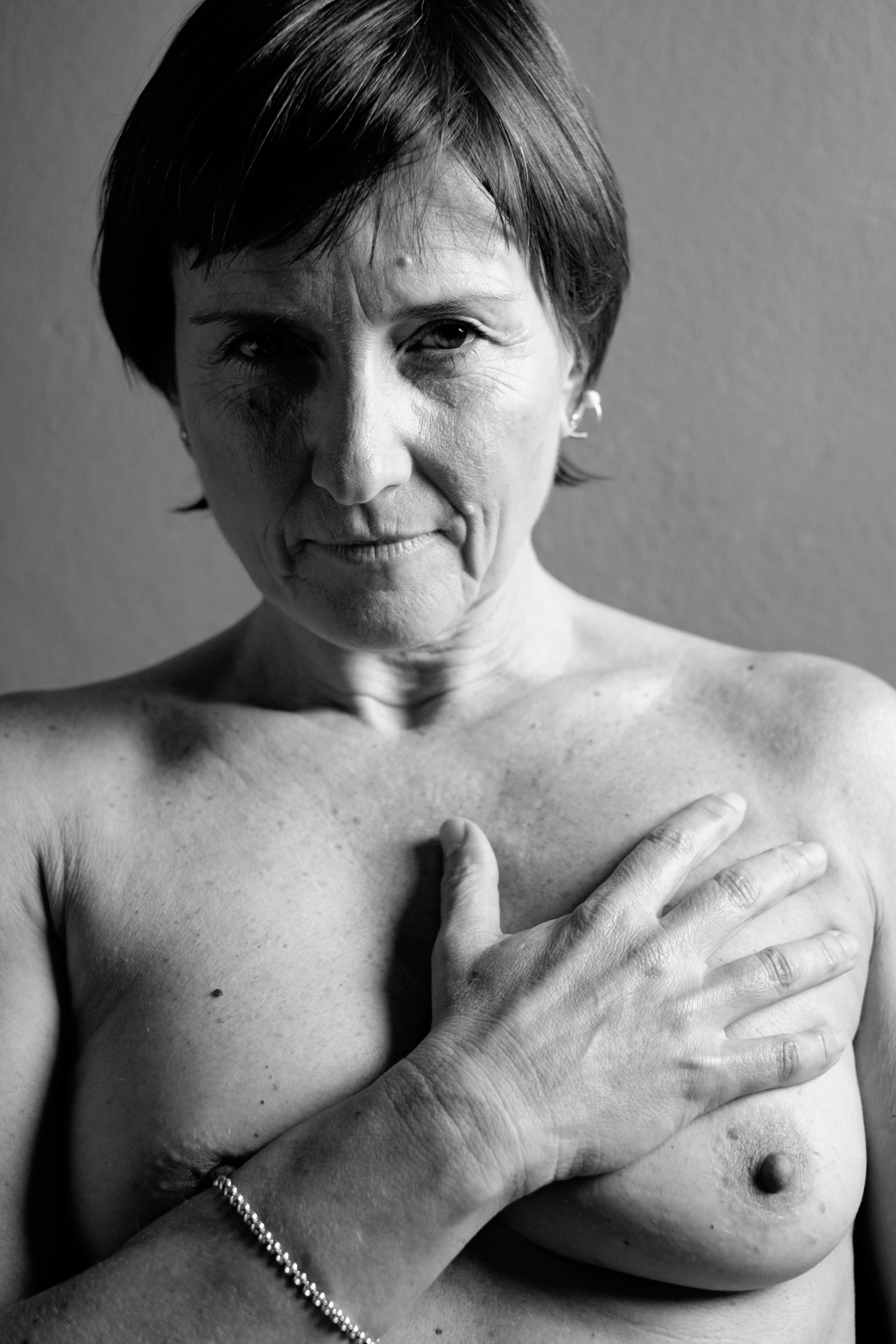 Mezza figura frontale a torace nudo; donna con capelli corti guarda in camera, mano destra appoggiata al petto, pelle segnata, luce morbida.