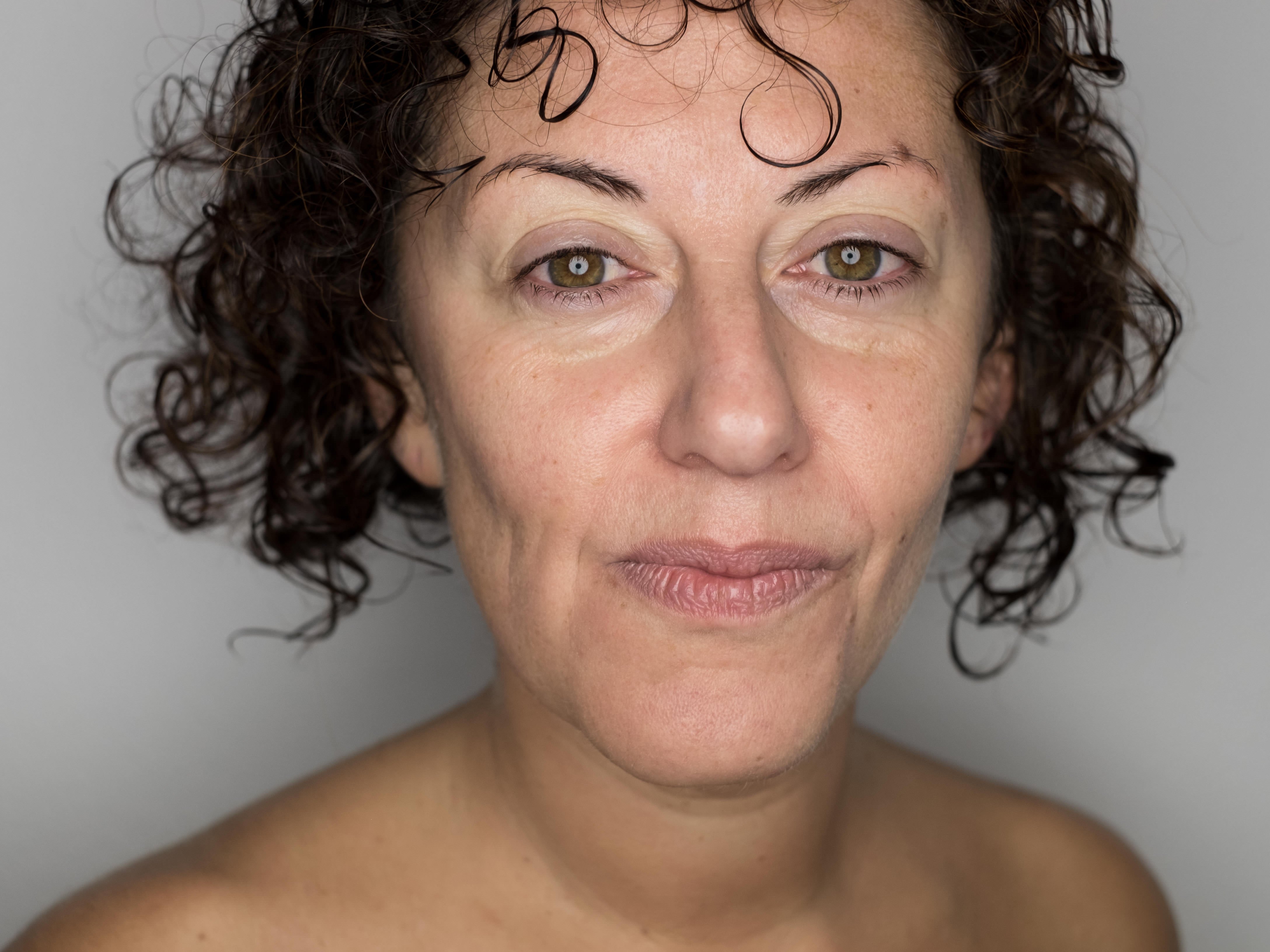 Primo piano a colori del volto di una donna con capelli corti e ricci; sguardo diretto verso la fotocamera, espressione neutra con labbra chiuse, linee sottili e piccole rughe visibili intorno agli occhi; inquadratura che mostra il viso fino alle spalle, luce frontale morbida che modella i lineamenti e sfondo chiaro uniforme senza elementi di distrazione.