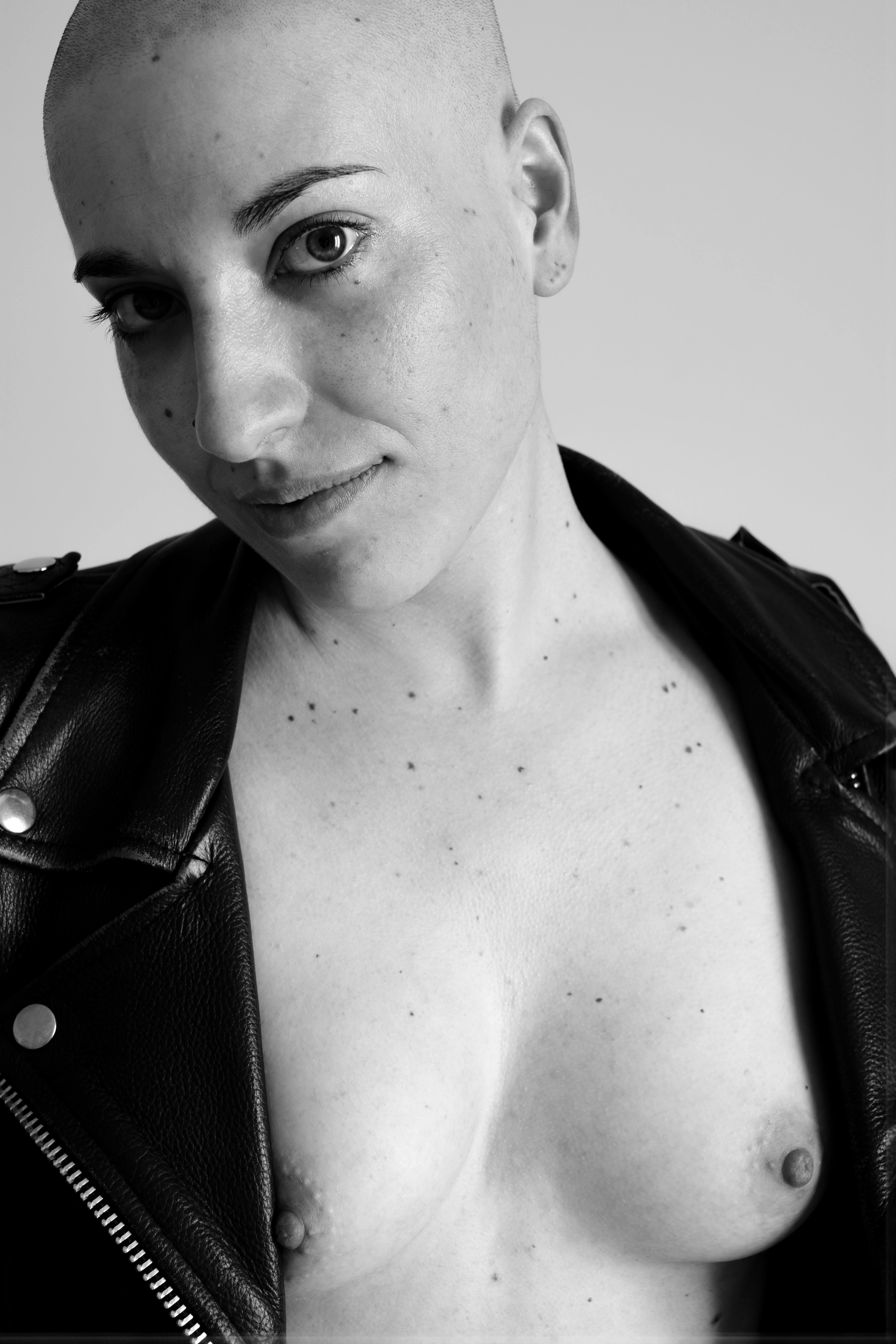 Ritratto in bianco e nero di donna con testa rasata che indossa una giacca di pelle aperta; busto esposto fino al petto, seni visibili, espressione diretta e confidenza nello sguardo verso l’obiettivo; pelle chiara con piccole macchie o nei sul petto, luce frontale morbida che riduce le ombre sul volto.
