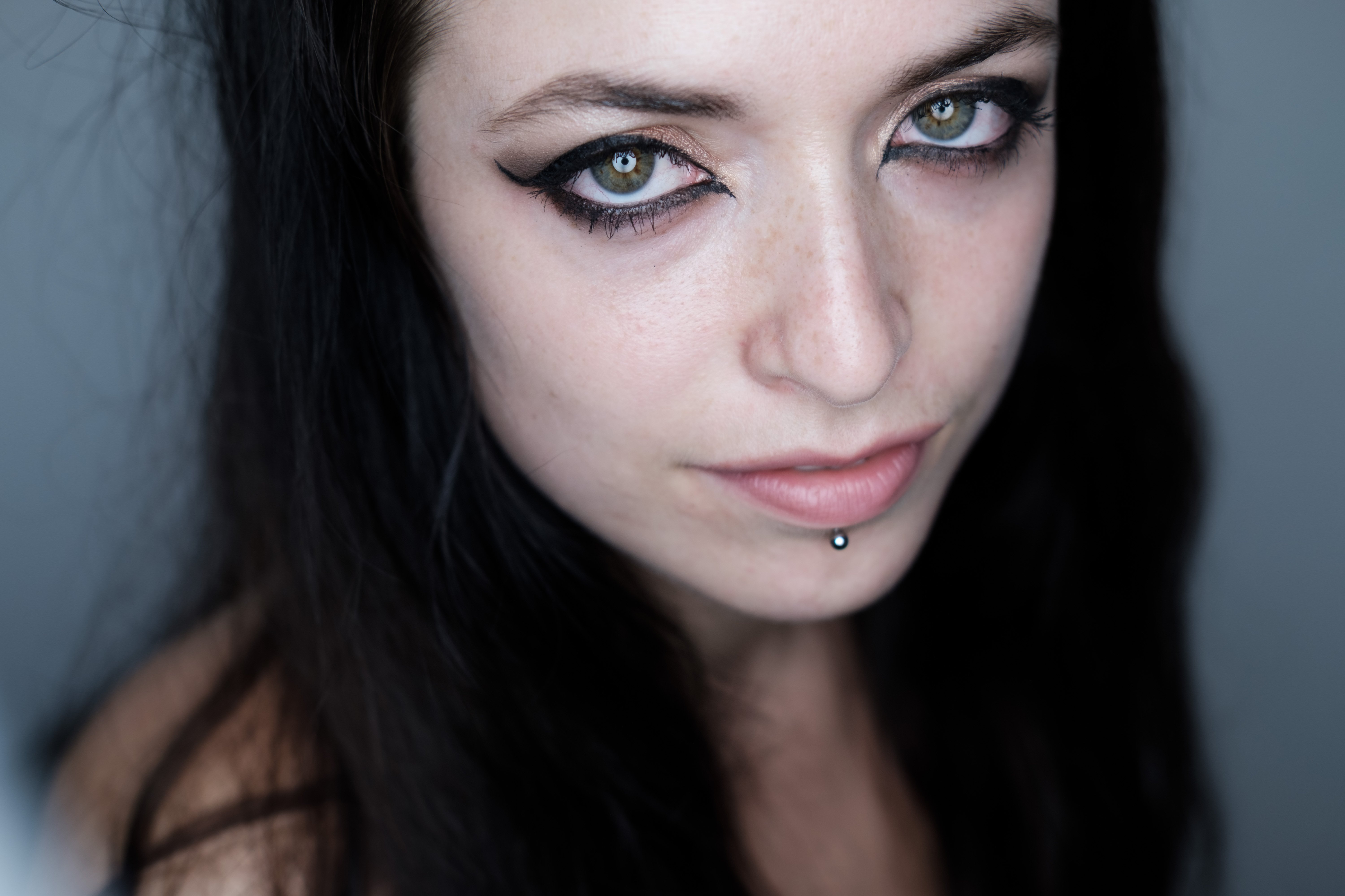 Primo piano a colori del volto con luce naturale; donna con capelli scuri lunghi, sguardo diretto alla fotocamera, pelle uniforme e espressione neutra; inquadratura mostra il volto fino alle spalle, sfondo esterno sfocato.