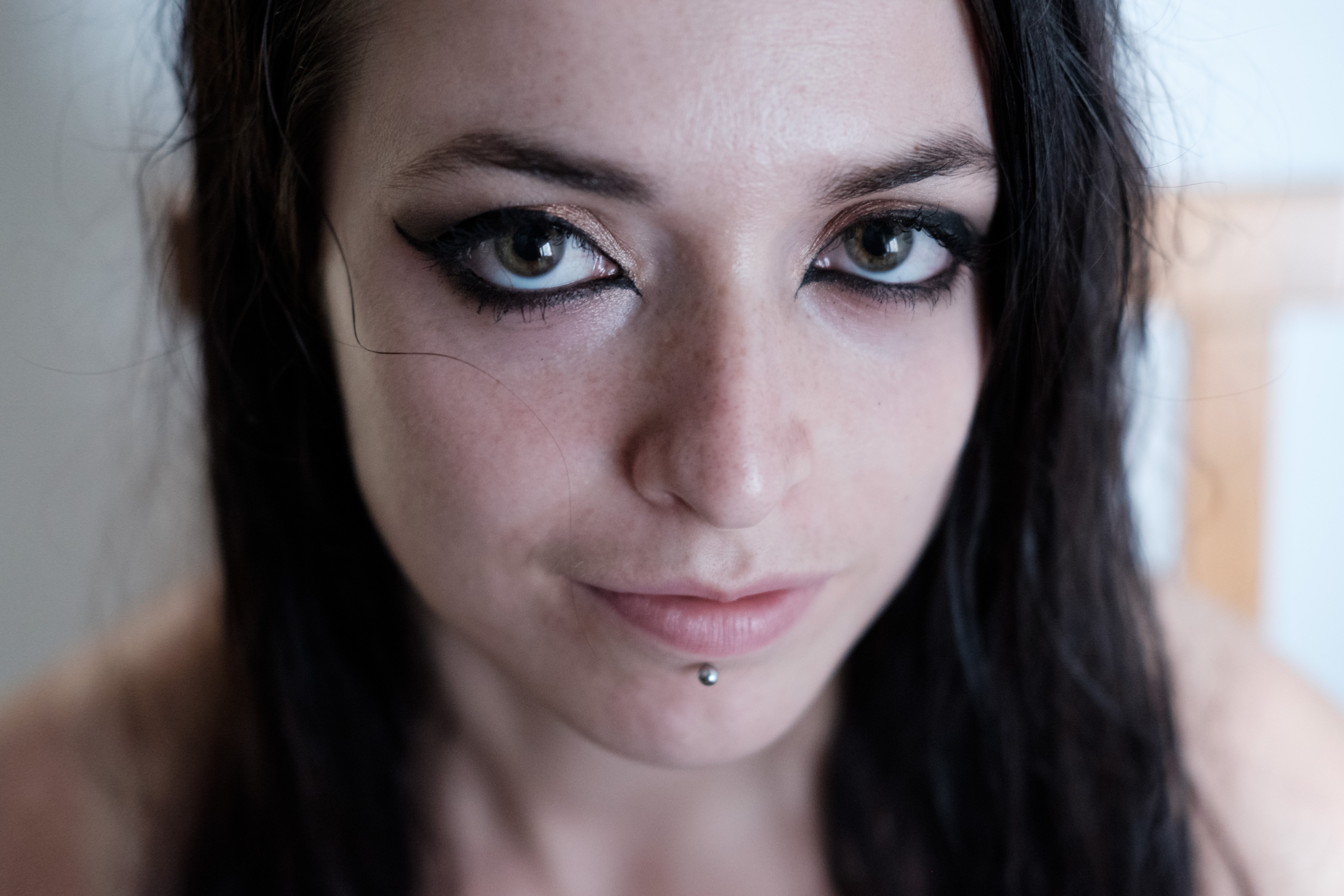 Primo piano a colori del volto con trucco leggero; occhi evidenziati da eyeliner scuro, labbro inferiore trafitto da un piercing, espressione neutra, capelli scuri lisci sulle spalle; sfondo interno sfocato.