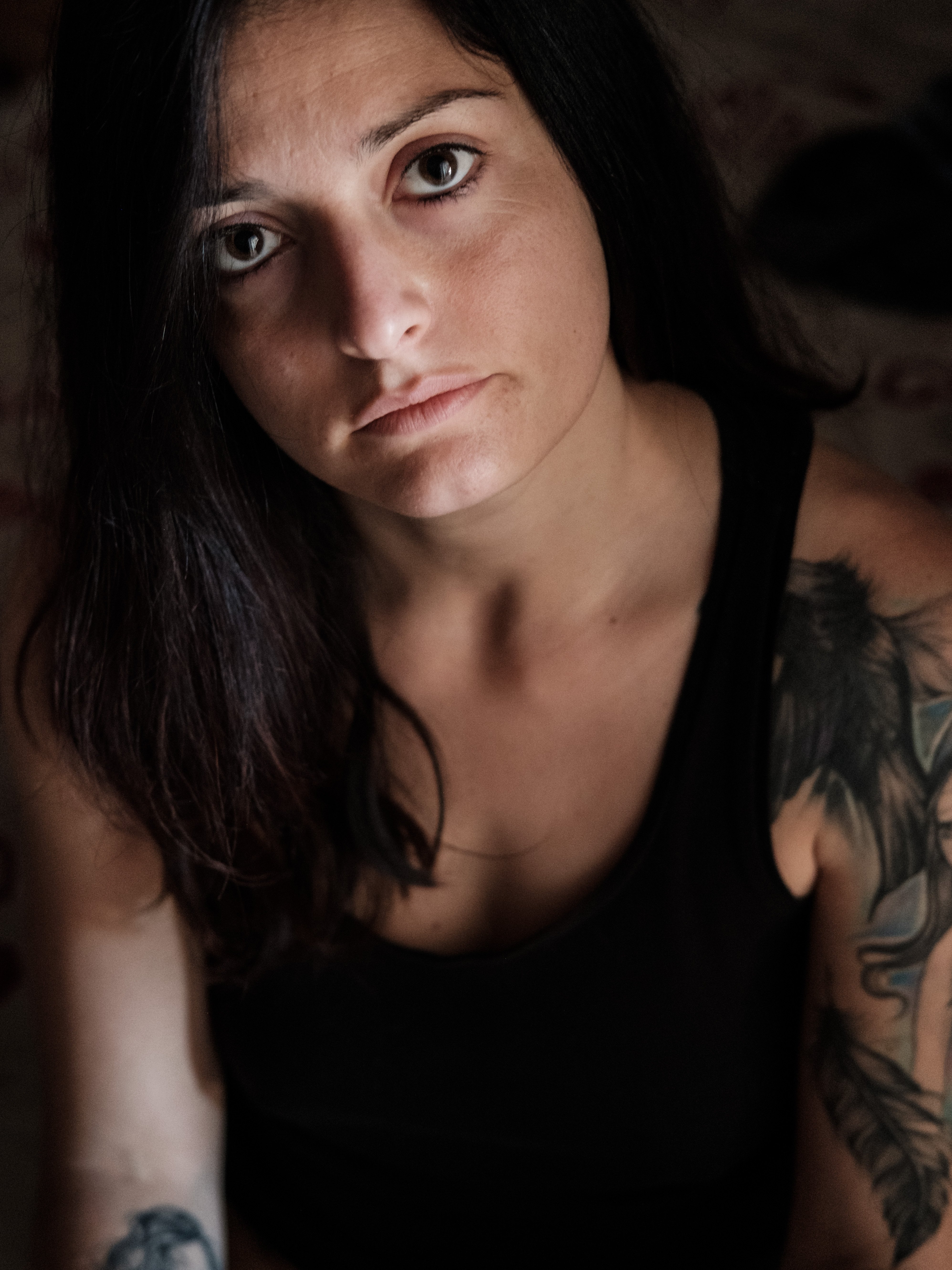 Primo piano a colori di una giovane donna con occhi scuri e trucco marcato; sguardo diretto verso l'obiettivo, labbro inferiore trafitto da un piercing, capelli lunghi scuri; luce naturale morbida, sfondo interno sfocato.