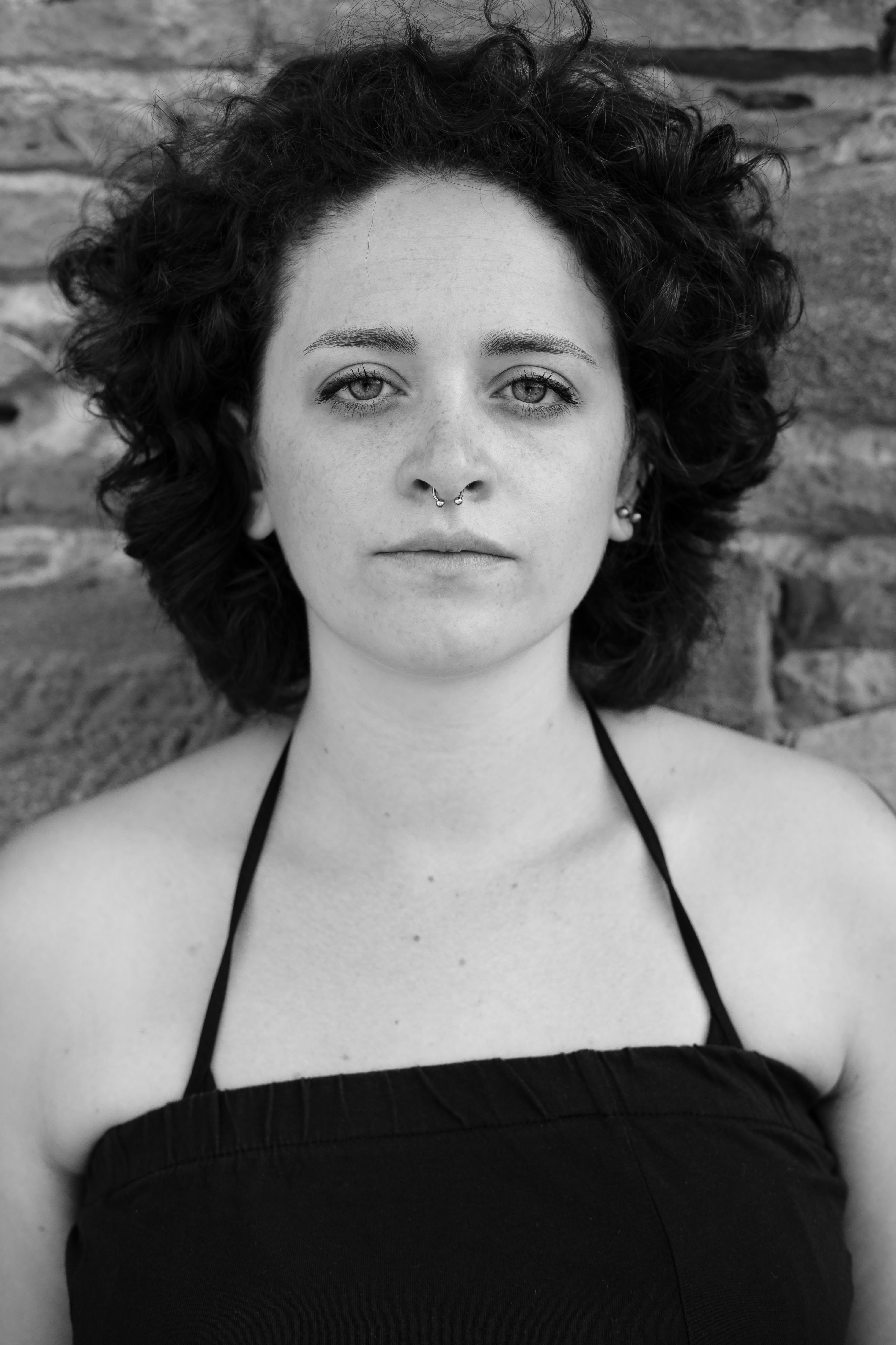 Primo piano in bianco e nero di una donna con capelli corti e ricci; occhi aperti, volto rilassato, indossa un top nero con spalline sottili; luce frontale morbida, sfondo in pietra sfocato.